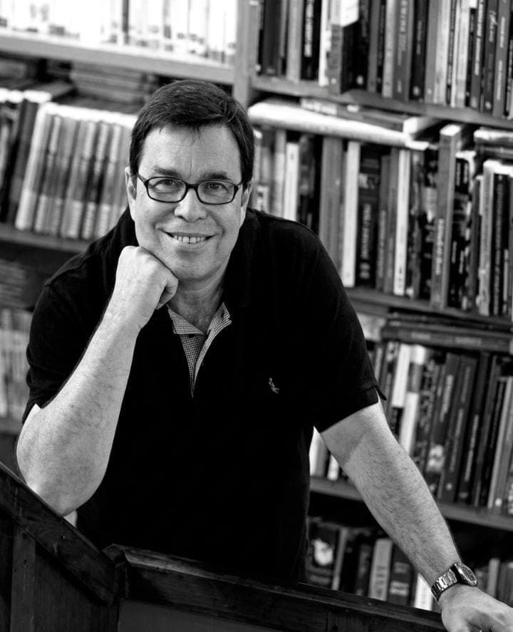 Retrato em preto e branco de Roberto Halfin, de óculos, apoiado em um corrimão diante de uma estante de livros